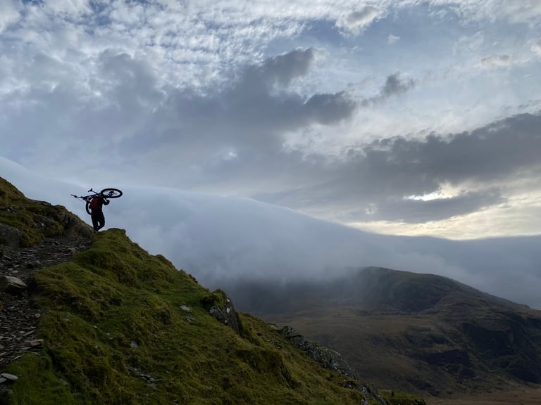 Silhouetted rider carries a mountain bike on their shoulder along a grassy ridge above cloud on Snowdon.