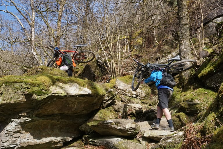 Two riders shoulder their mountain bikes up mossy rock steps in woodland on the path to Twm Sion Cati's Cave.
