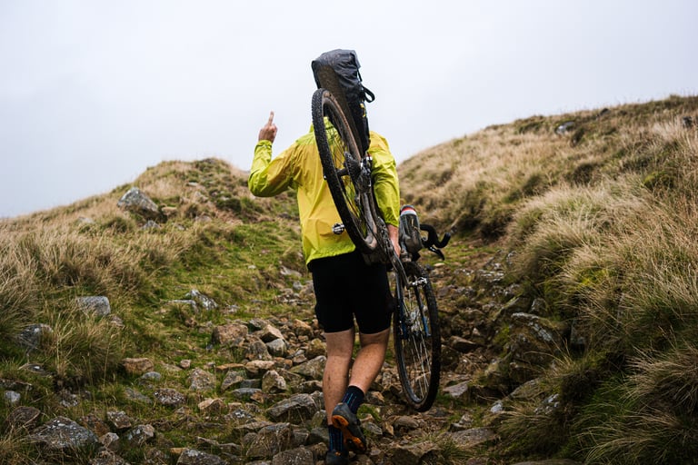 Rider in a yellow jacket shoulders a loaded gravel bike up a rocky, grassy hillside during hike-a-bike.