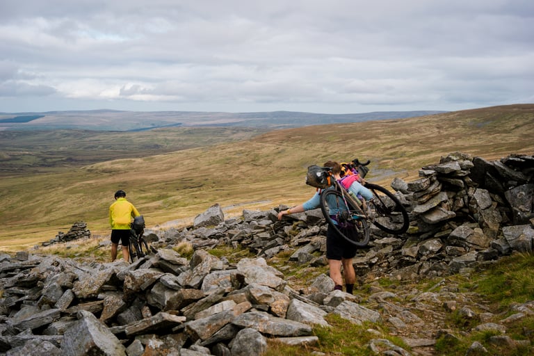 Rider shoulders a bikepacking bike across a boulder-strewn moor while a companion in a yellow jacket walks ahead.
