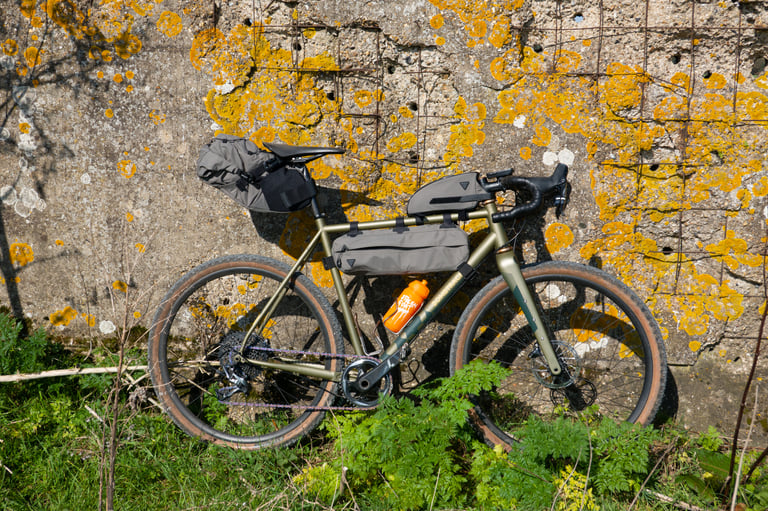 Green Bombtrack loaded with the full Topeak bikepacking set and a bottle, propped against a lichen-spotted wall.