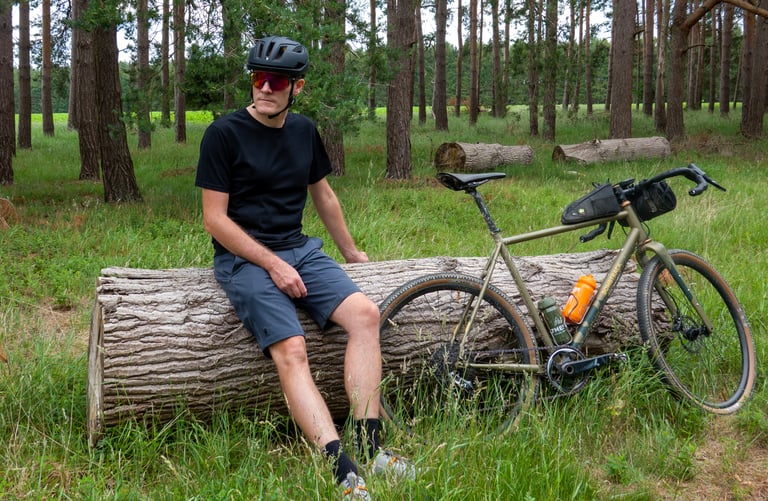 Rider in Folsom shorts and black tee sits on a fallen log in pine woodland beside a gravel bike.
