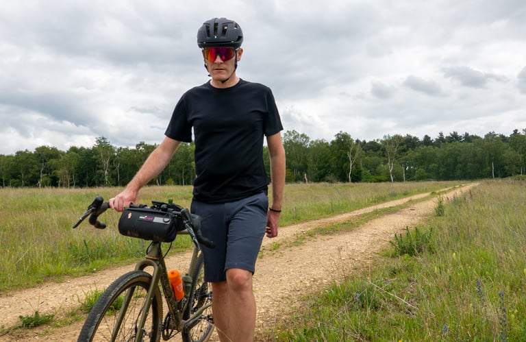 Rider in the Folsom Short 2.0 walks a gravel bike with a bar bag along a dirt track through open grassland.