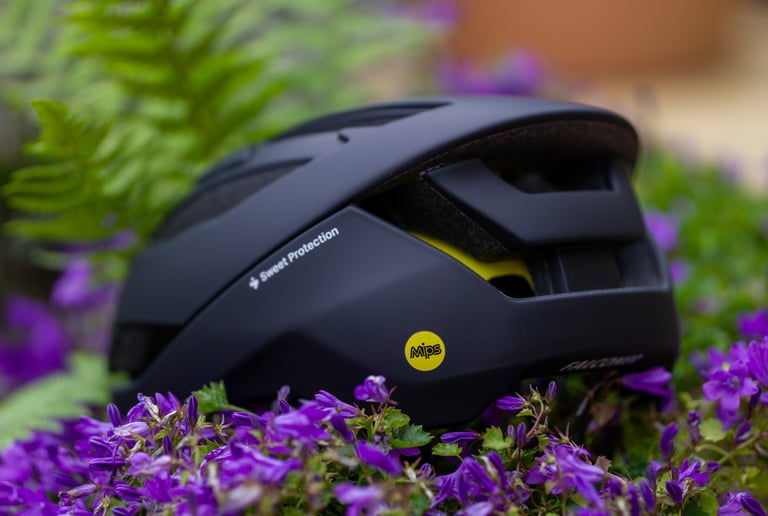 Side of the matte black Sweet Protection Falconer II helmet, with its yellow MIPS badge, among purple flowers.