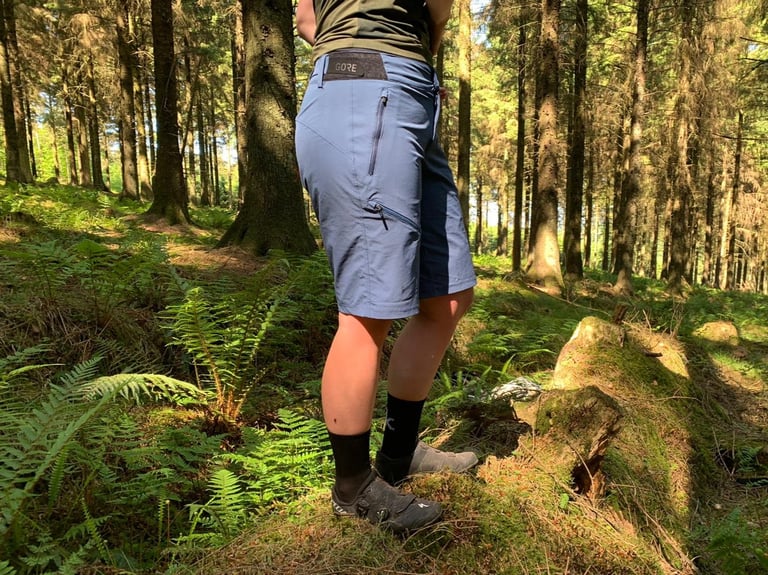 Rear three-quarter view of the GORE C5 Women's Shorts worn in a forest, with a zipped hip pocket and GORE waist tab.