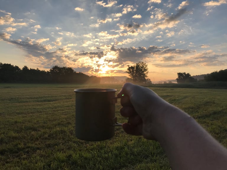 Hand holding a camping mug of coffee at sunrise over a misty field on the solstice morning of the Grenzsteintrophy.