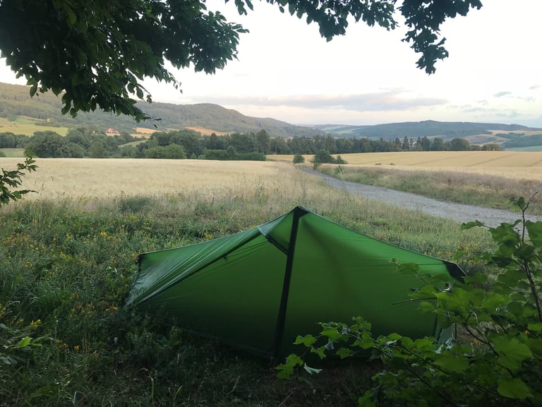 Green one-person tent pitched in a field above rolling hills at dusk, a Grenzsteintrophy wild camp.