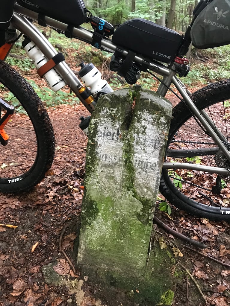 Mossy stone border marker on a forest trail beside a loaded Kinesis bikepacking bike with Apidura bags.