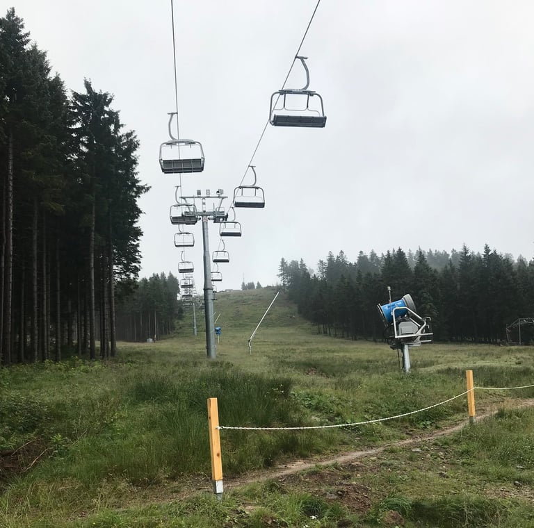 Empty chairlift and a blue snow cannon on a grassy Brocken ski slope under grey skies, Harz mountains.