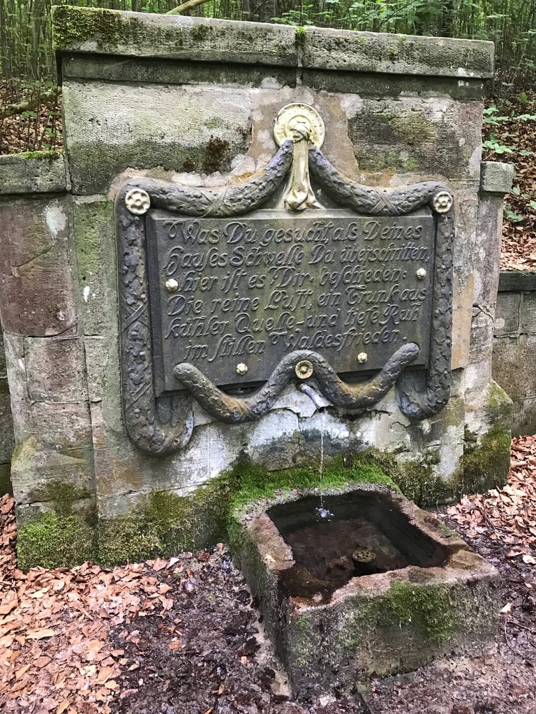 Moss-covered stone fountain at Rödental with an ornate cast inscription above the spout, on the Grenzsteintrophy route.