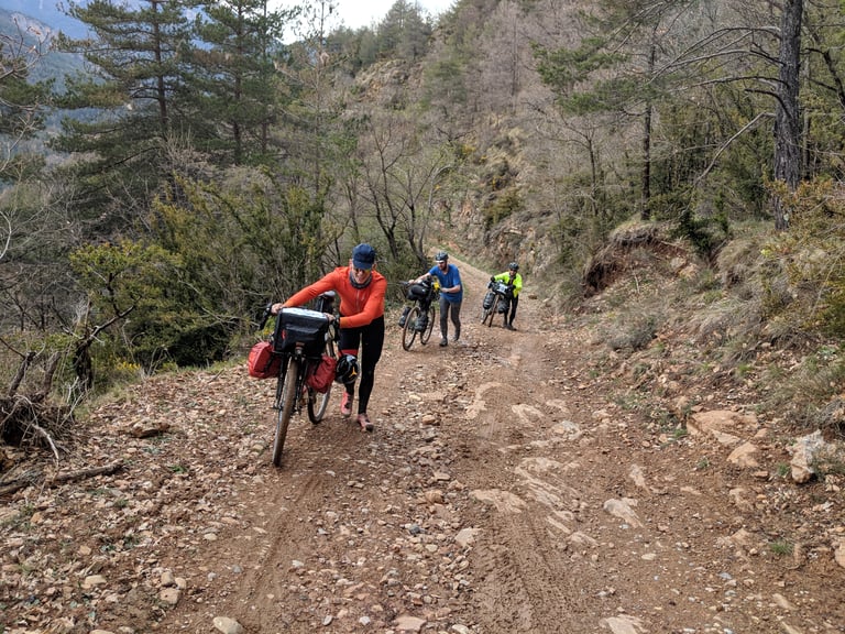 Three bikepackers push loaded bikes up a steep, loose track through scrub in the Pyrenees.