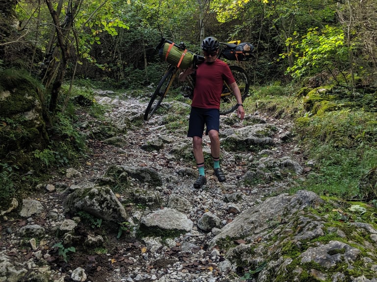 Bikepacker in a red top shoulders a loaded bike, cyclo-cross style, along a rocky wooded track.