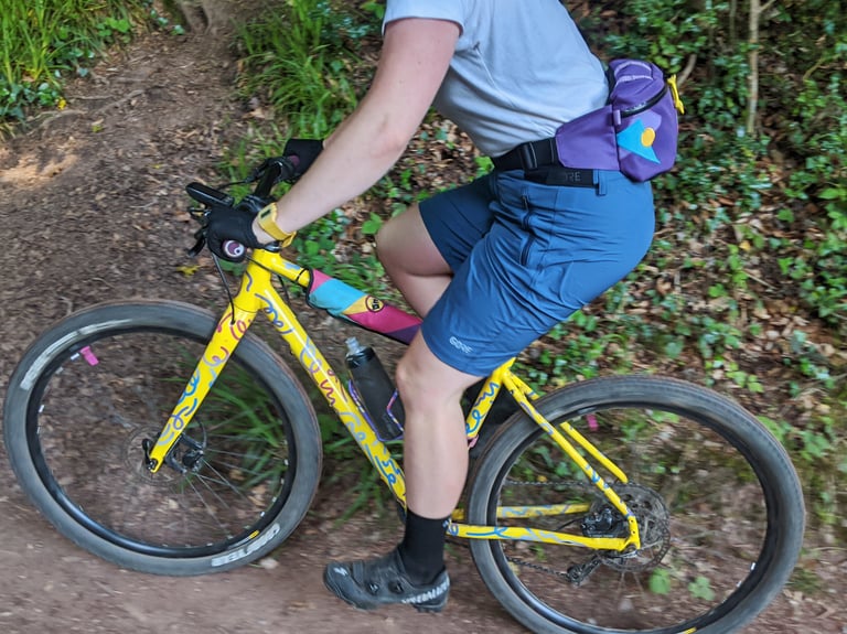 Rider in GORE C5 Women's Shorts on a yellow gravel bike with a purple bum bag, riding a dirt trail.