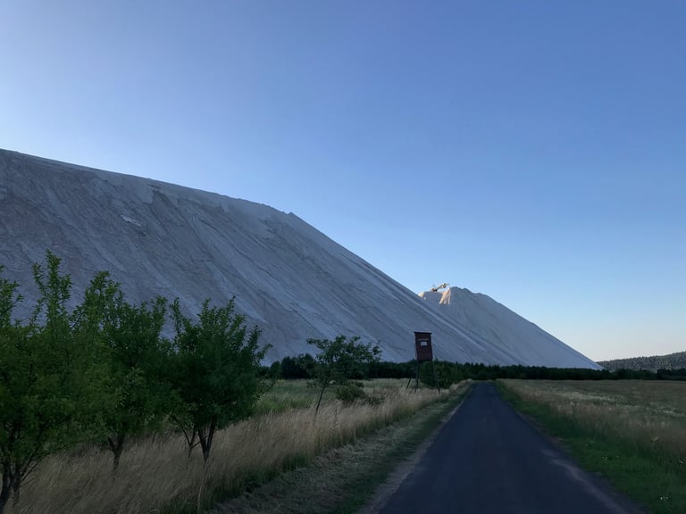 Vast white salt heap rising above a tarmac road and fields, with a machine on its summit, near Vacha.
