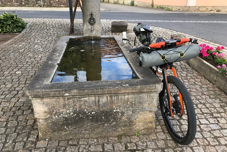 Kinesis mountain bike with Apidura bags and orange Jones bars leant against a stone village fountain in Germany.