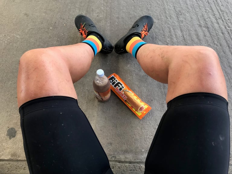 Rider's legs and cycling shoes stretched out on concrete beside a BiFi Roll XXL sausage snack and a small bottle of drink.