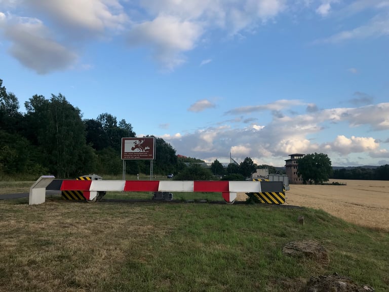 Red and white steel ram barrier at Teistungen border control point with a watchtower and information sign behind.