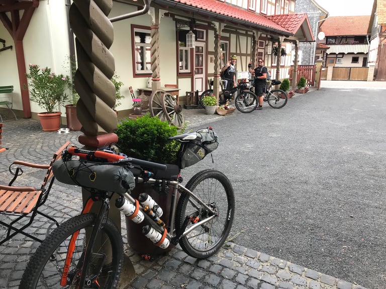 Two bikepackers wave in a village courtyard of half-timbered buildings, with a loaded mountain bike in front.