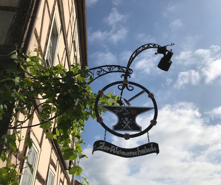 Wrought-iron pub sign reading Zur Weimarschmiede with an anvil, hanging from a half-timbered building in Germany.