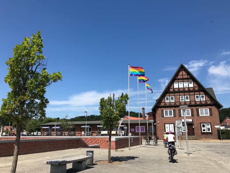 Rainbow flags flying in a small German town square as a cyclist rides towards a half-timbered brick building.