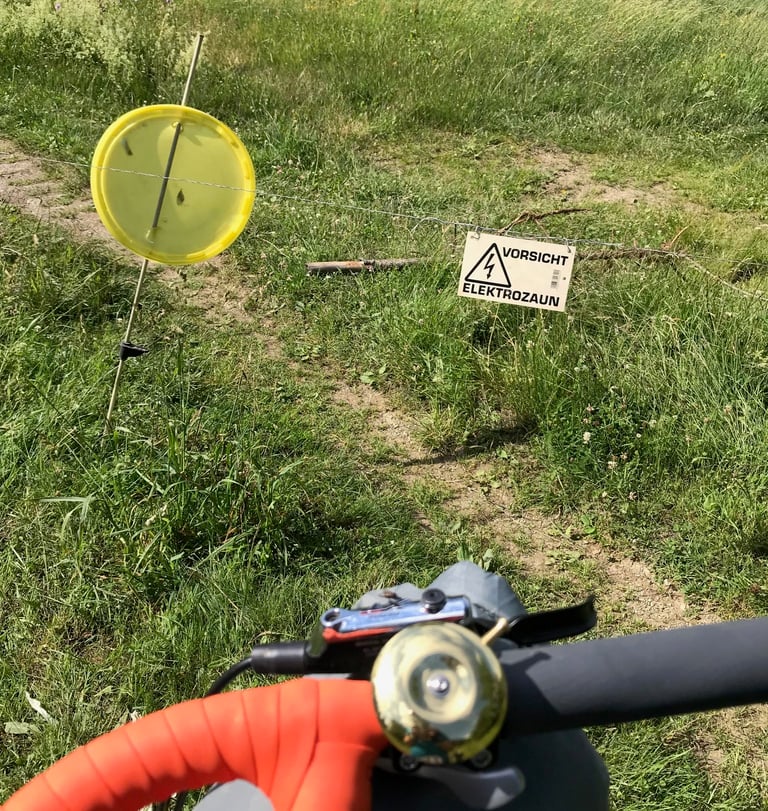Electric fence across the track with a yellow marker and Vorsicht Elektrozaun sign, seen over the handlebars.