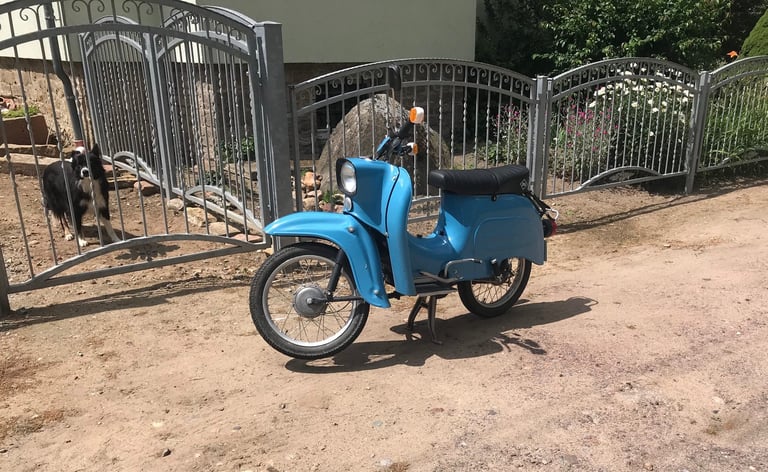 Blue East German Simson scooter parked outside a metal gate, a dog watching from behind it.