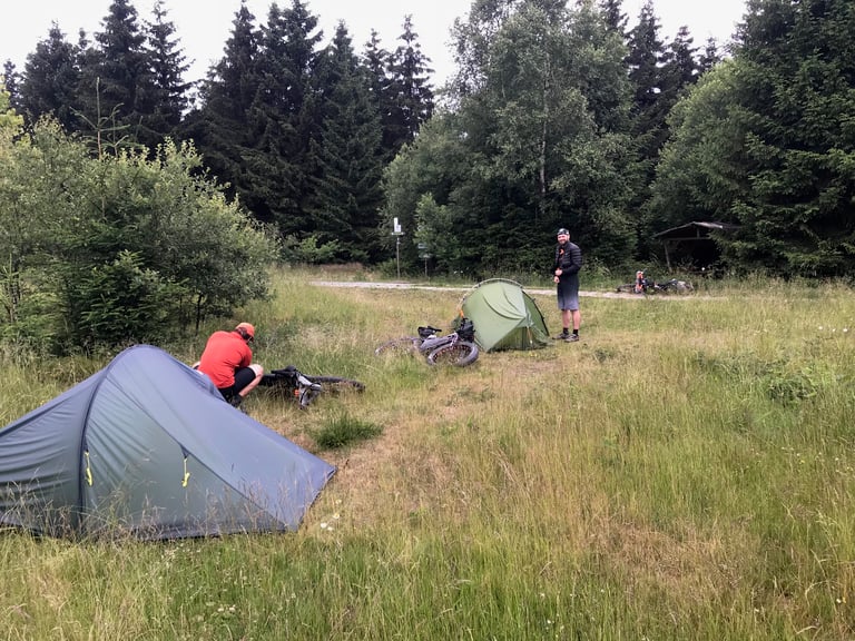 Two bikepackers break camp beside their tents in a grassy forest clearing on the Grenzsteintrophy route in Germany.