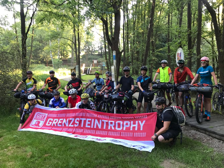 Grenzsteintrophy 2018 starters with loaded bikes and the GST banner in woodland at the Staatsgrenze start near Hof.