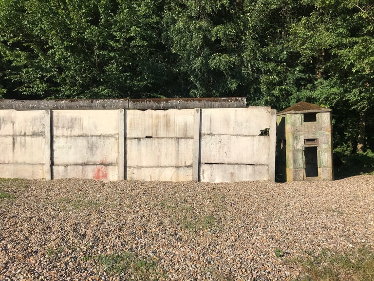 Preserved section of concrete border wall beside a small concrete hut, gravel in front and woods behind.