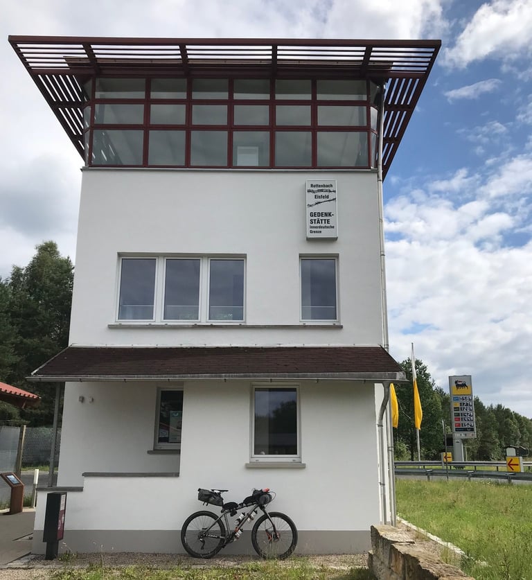 Restored Inner German border watchtower beside a motorway, with a loaded bike leaning against it on the Grenzsteintrophy.