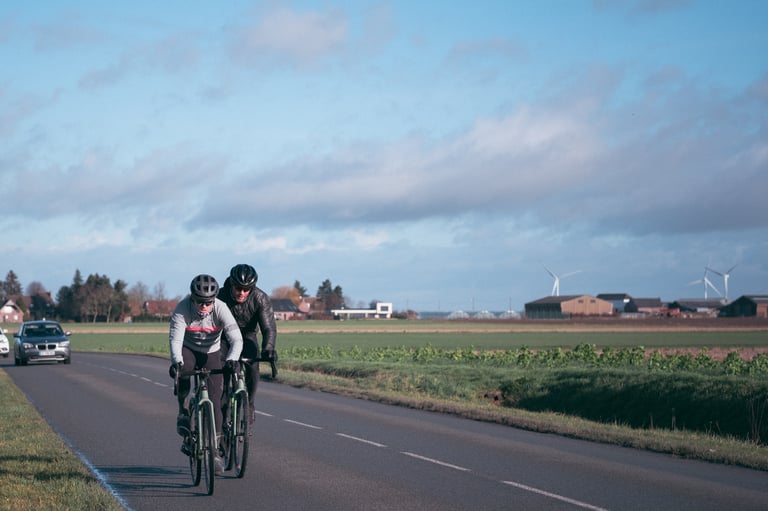Two riders in helmets ride side by side on a quiet Flemish road past fields, on the Santa Cruz Belgium road trip.