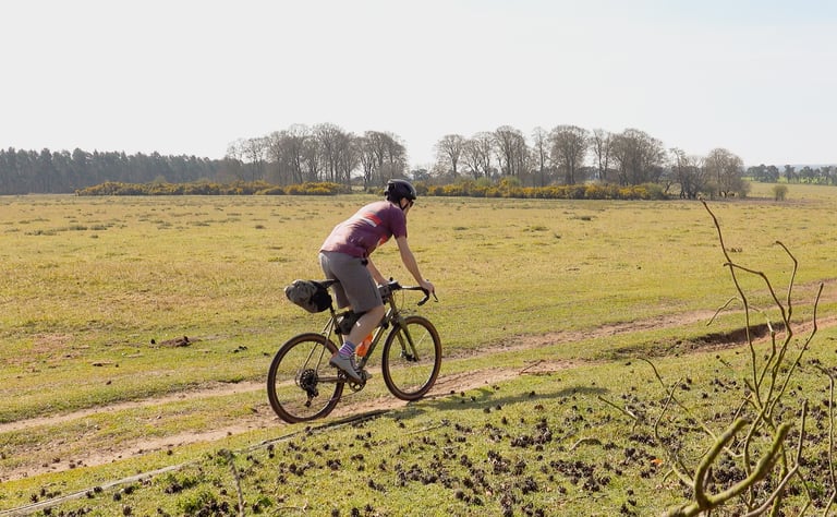 Rider on a Bombtrack gravel bike with a Topeak saddle bag follows a dirt track across open grassland.