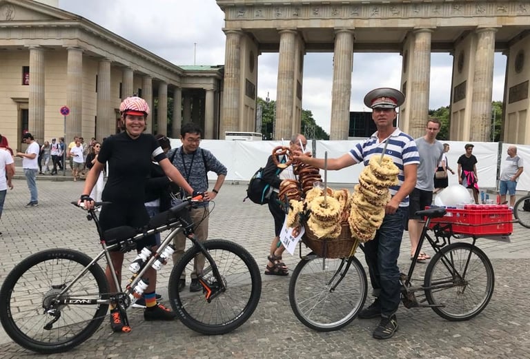 Rider with a mountain bike beside a pretzel seller in a peaked cap, in front of the Brandenburg Gate in Berlin.