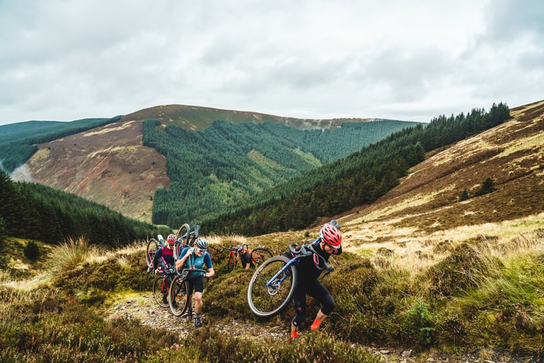 Frontier 300 riders shoulder their gravel bikes on the hike-a-bike climb over heather moorland above forest.