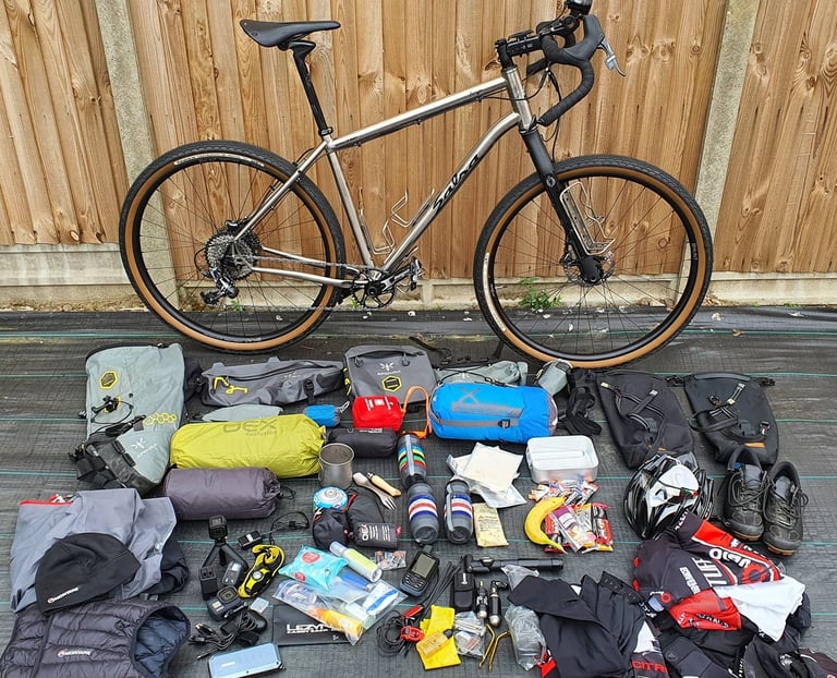 Salsa Fargo Ti against a wooden fence, with its Apidura bags, tent, sleeping bag, helmet and snacks laid out below.
