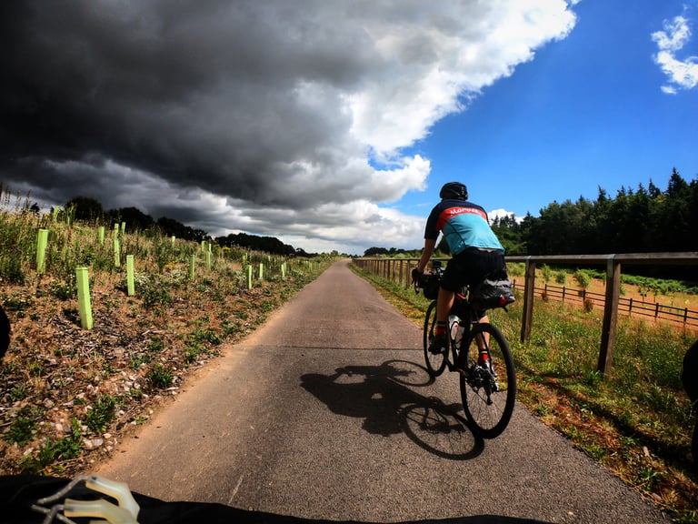 Rider on a narrow tarmac path in Norfolk heads towards a dark rain cloud, while blue sky shows to the right.