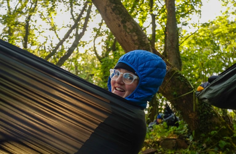 Author Katherine Moore grins from her hammock in a blue insulated hood, slung between trees in woodland.