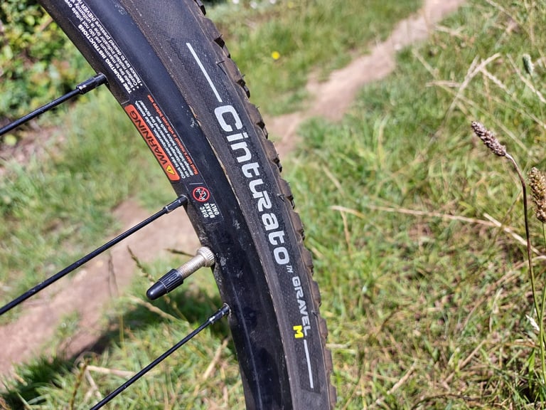 Close-up of the Pirelli Cinturato Gravel M sidewall on a wheel, with valve and spokes beside a grassy track.