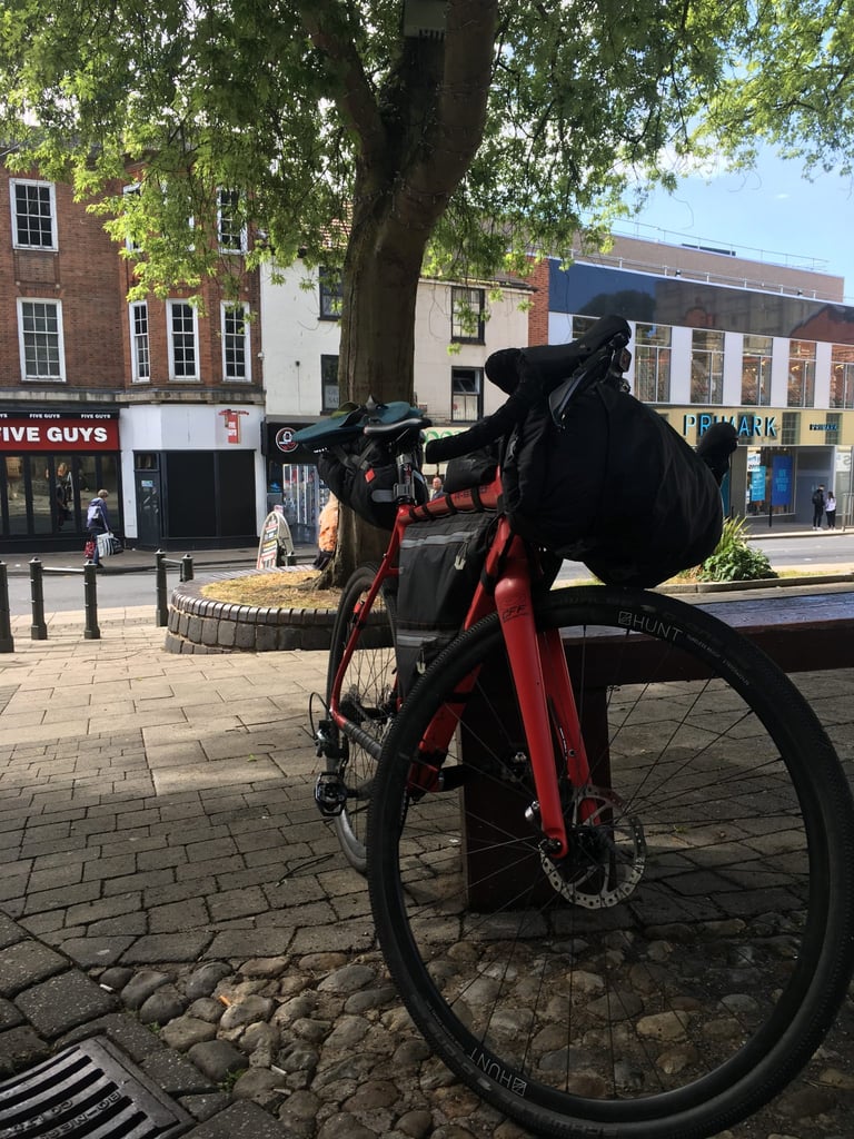Red loaded Thompson gravel bike on Hunt wheels leans on a bench under a tree in Norwich city centre at the ride's end.