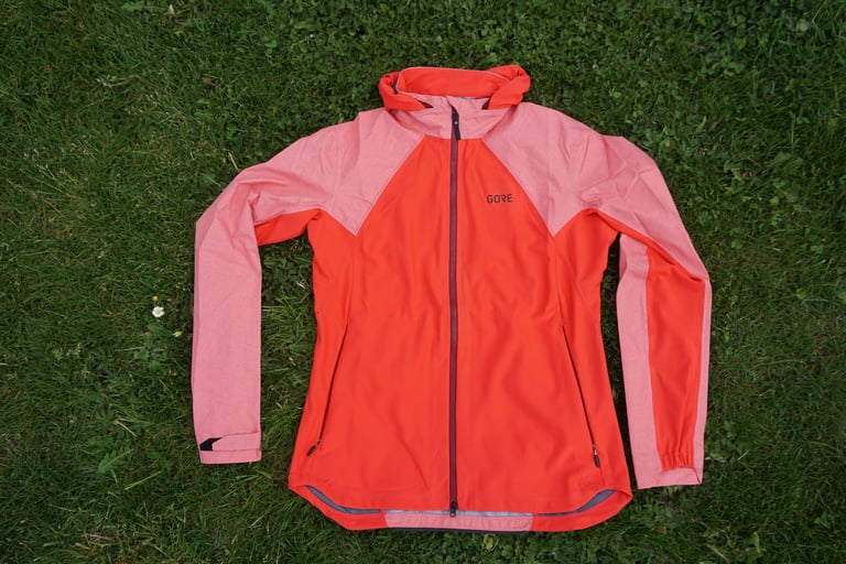 GORE Infinium Hybrid Jacket in hibiscus pink laid flat on grass, front view with two-way zip and hood rolled down.