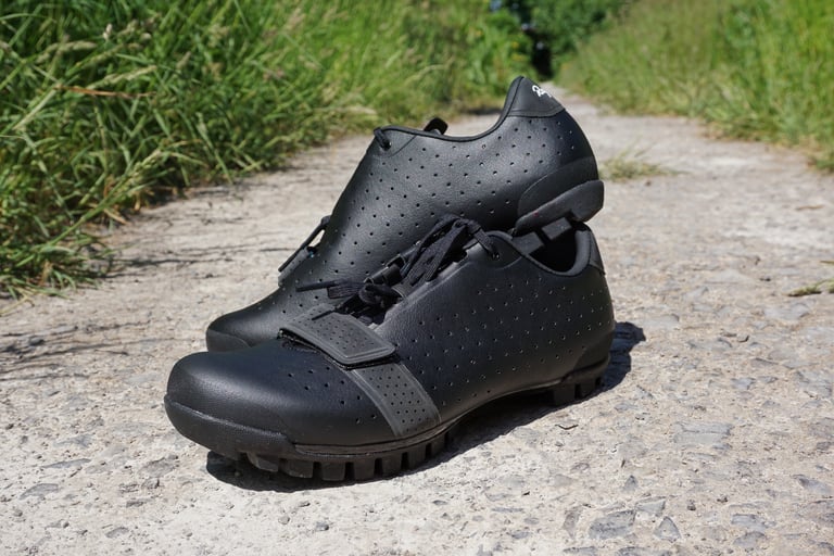 Black Rapha Explore Shoes resting one on the other on a grassy track, showing the classic leather-look styling.