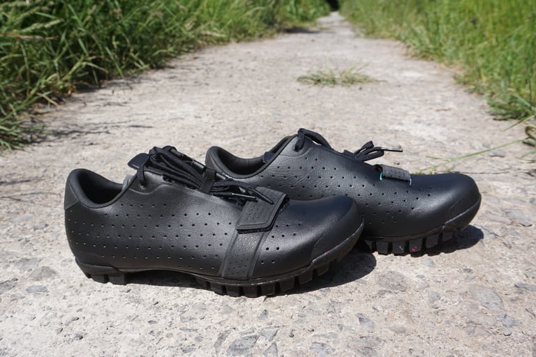 Pair of black Rapha Explore Shoes side by side on a stony path between grass, showing the high heel cup.