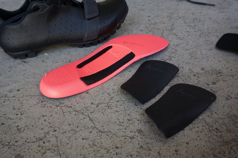 Pink insole and two black arch support inserts supplied with the Rapha Explore Shoes, beside a shoe.