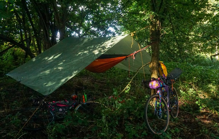 Orange hammock under a green tarp in woodland with two bikepacking bikes parked nearby, the morning after rain.