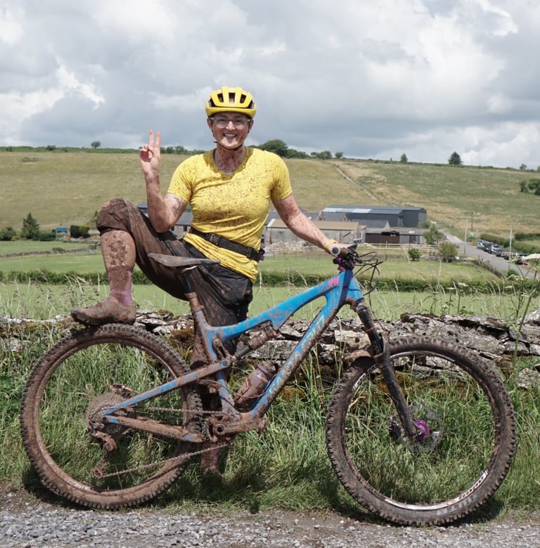 Mud-splattered Katherine in a yellow Velocio Radiator TRAIL tee gives a peace sign beside a mountain bike and stone wall.