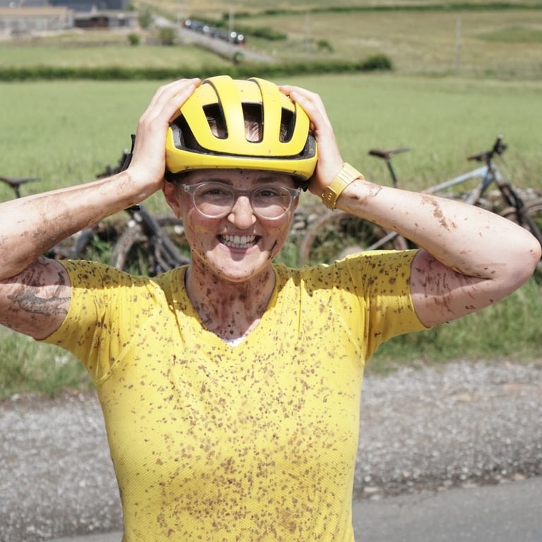 Katherine, splattered in mud in a yellow Velocio tee, grins with hands on her yellow helmet, bikes behind.