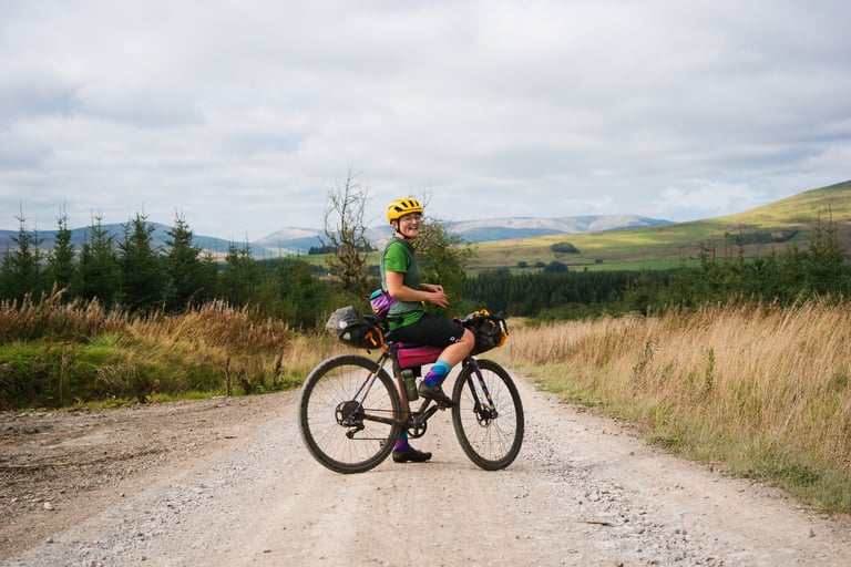 Katherine smiles over her shoulder astride her bikepacking-loaded Mercredi on a gravel road with hills behind.