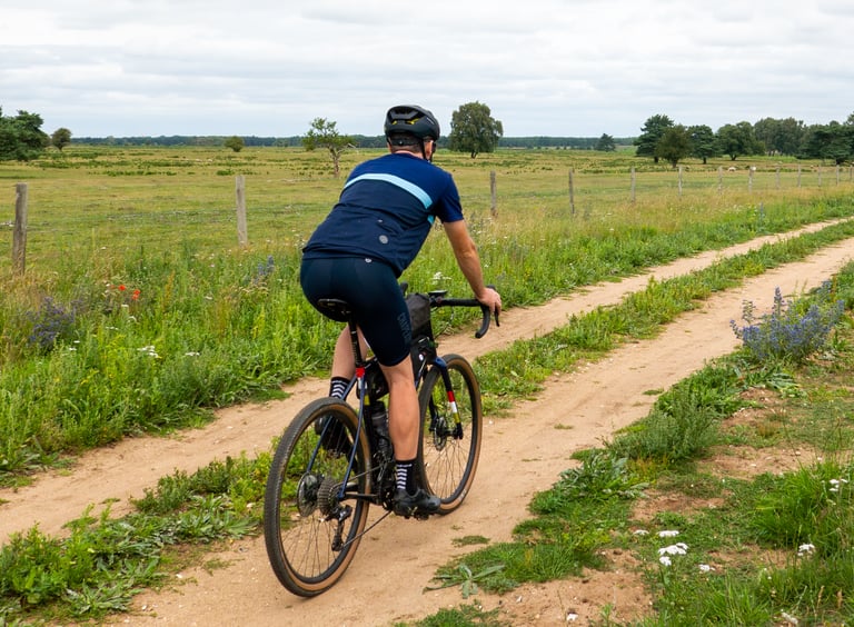 Rider in the Chapeau! Café jersey on a gravel bike along a sandy double-track past open Breckland pasture.