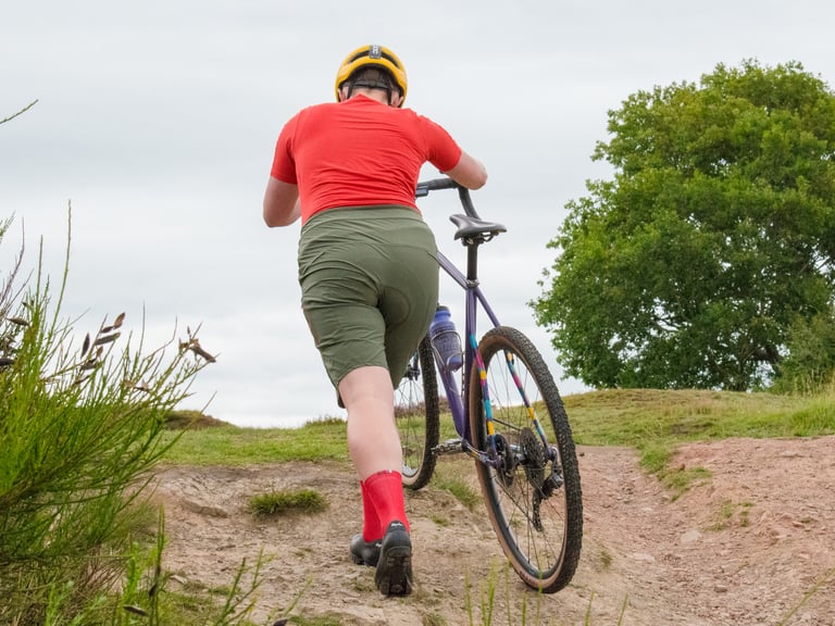 Rider in khaki Velocio TRAIL Shorts and red tee pushes a purple gravel bike up a stony slope, seen from behind.