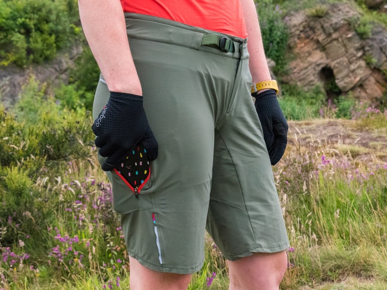 Velocio women's TRAIL Short in khaki with a hand in the zipped thigh pocket and a reflective strip on the leg.